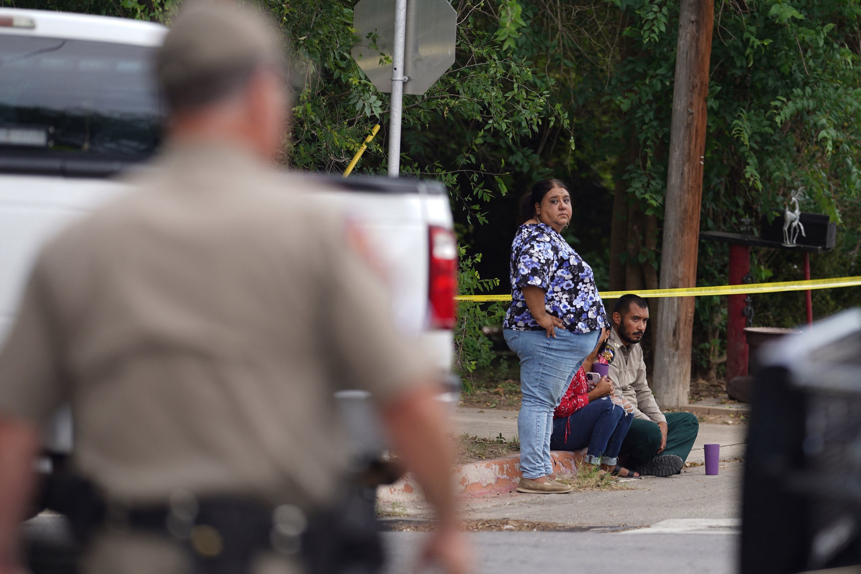 A woman in the background, standing by two people sitting on a sidewalk near some strung-up caution tape, looks toward a police officer in the foreground
