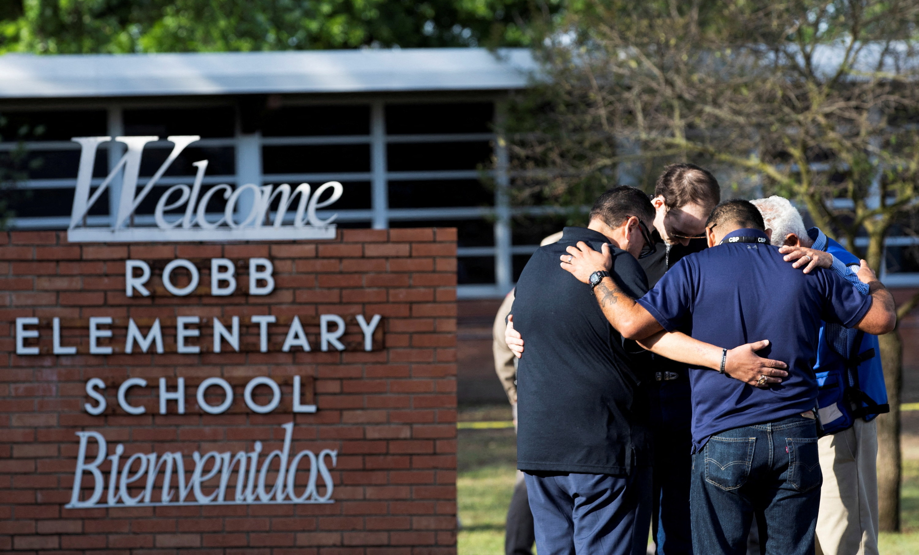 A group of people stand in a tight prayer circle beside the Robb Elementary welcome sign, with hands on each other&#x27;s backs and their heads bowed