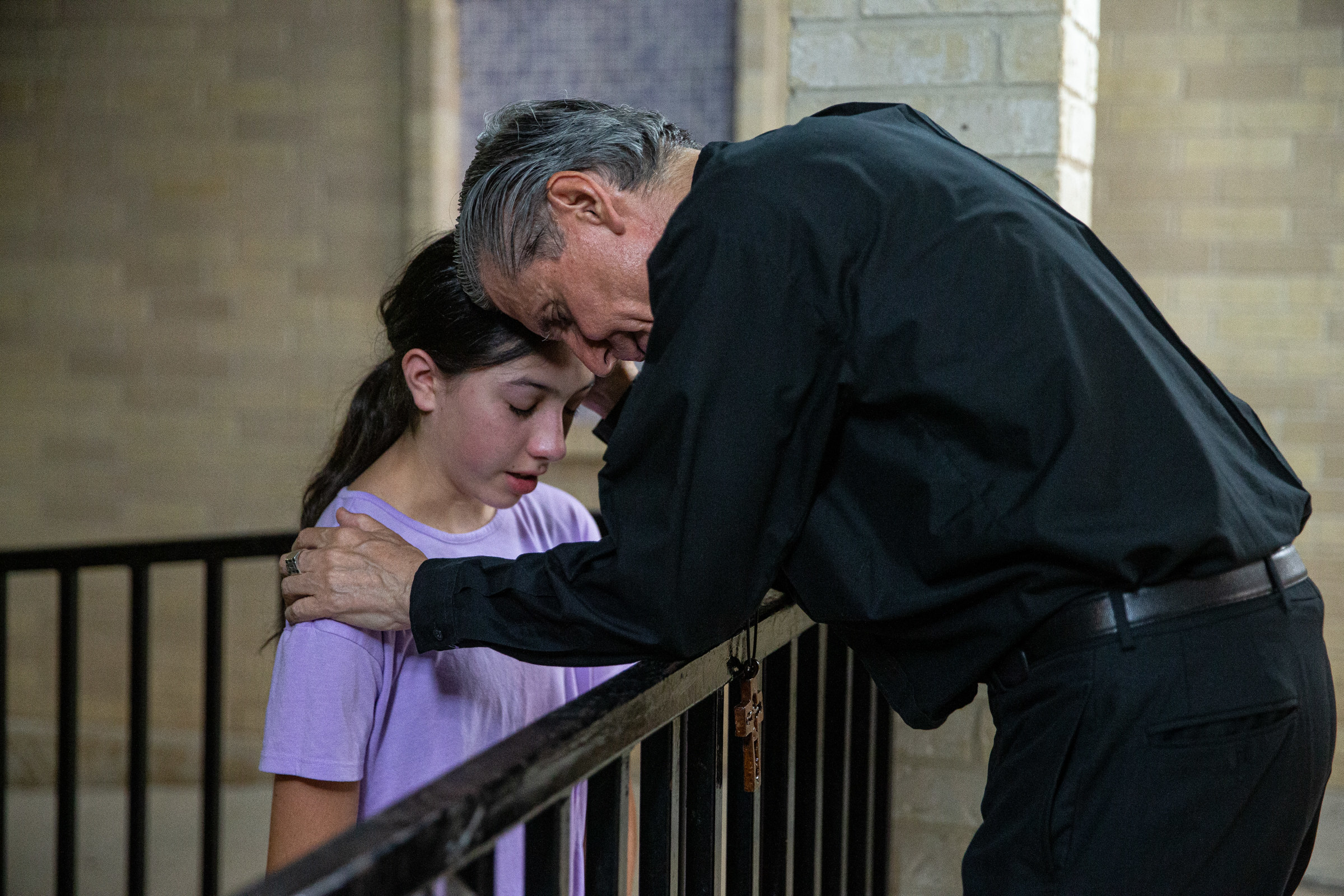 A man wearing all black leans over a gate and holds the shoulder of a young girl, as both pray