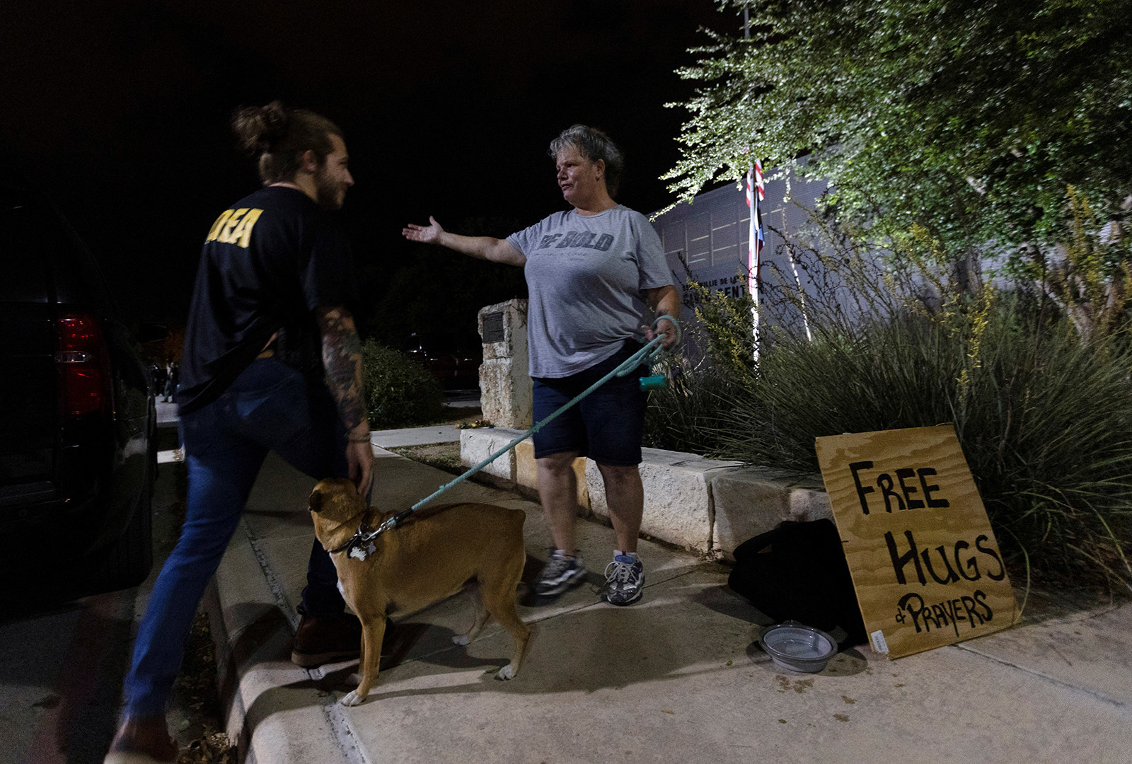 A man walks up to a woman who holds a dog on a leash and stands by a sign that reads &quot;free hugs and prayers&quot;