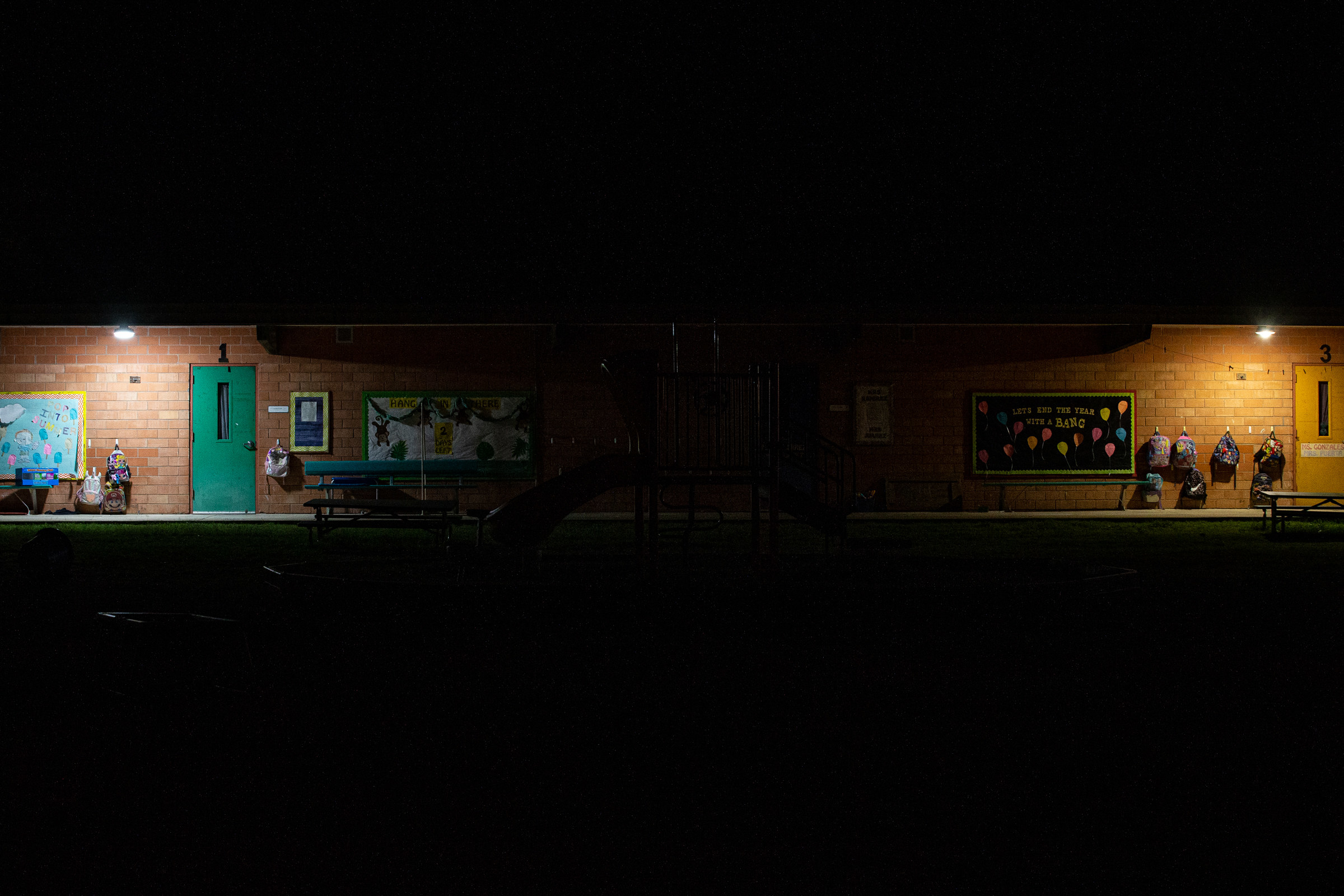 Overhead lights show classroom doors surrounded by brick walls and backpacks hanging on hooks overnight at a school