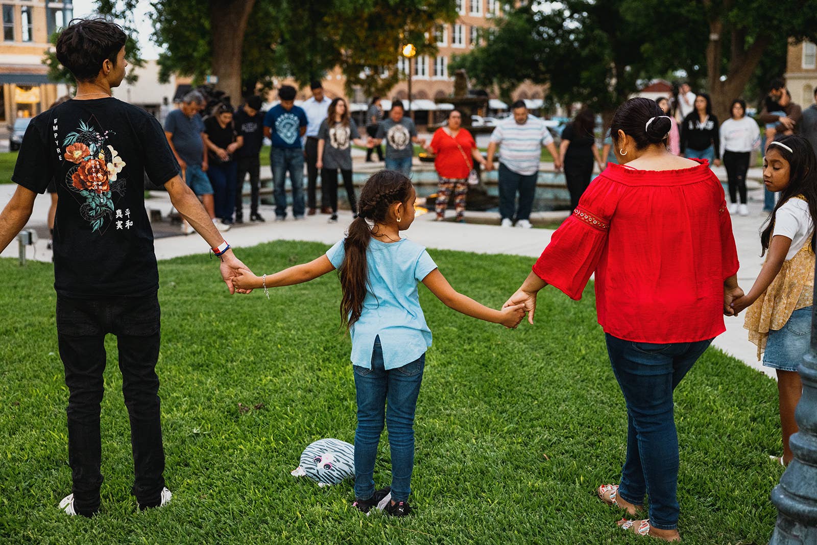 Adults and children stand in a large circle and hold hands outside on a plot of grass 