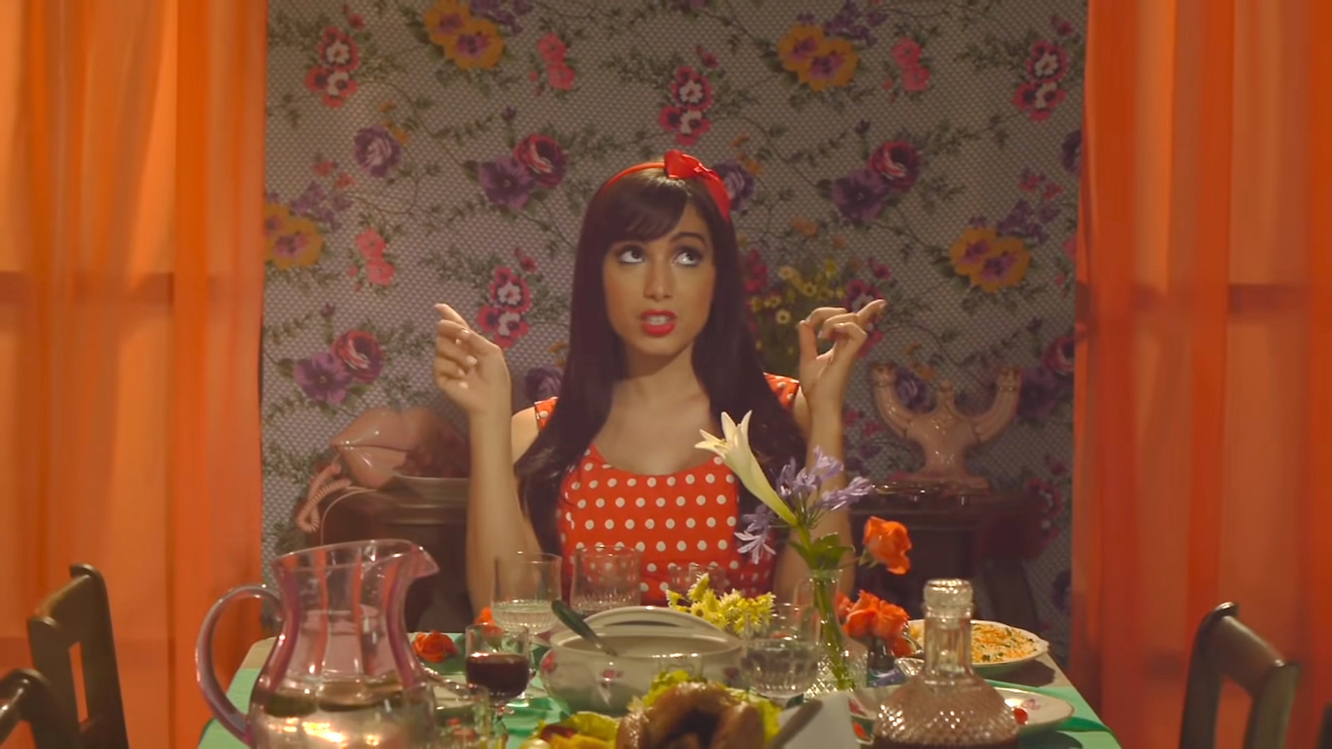 Anitta sitting at a table with lots of food and drink on it and looking up as she points upward