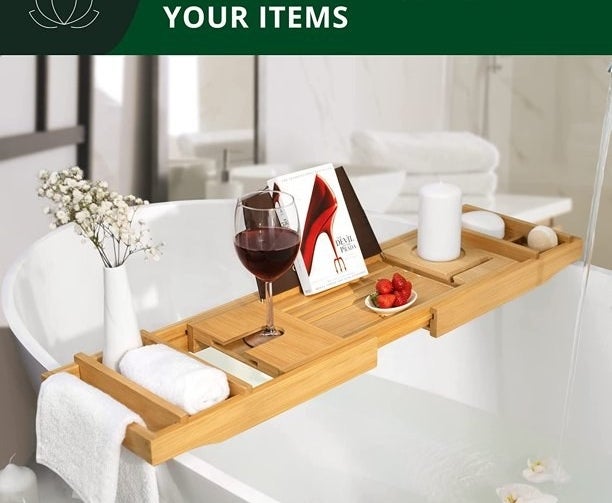 the bamboo bath caddy with towels, a book, a glass of wine, a bowl of strawberries, flowers and a candle on it