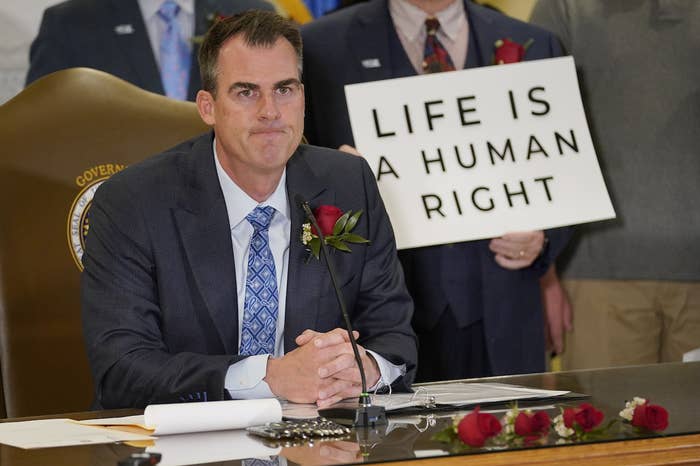 Kevin Stitt sits behind a desk near someone holding a sign reading &quot;life is a human right&quot;