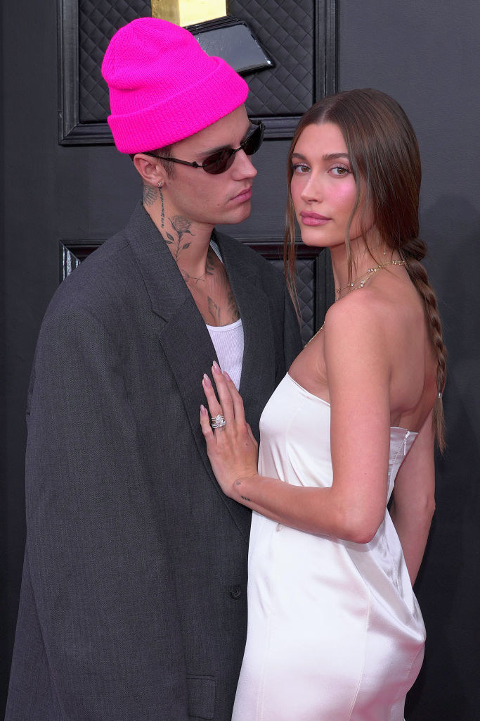 Justin, wearing a beanie and sunglasses, looking at Hailey, who&#x27;s wearing a slinky, strapless gown and has her hand on his chest