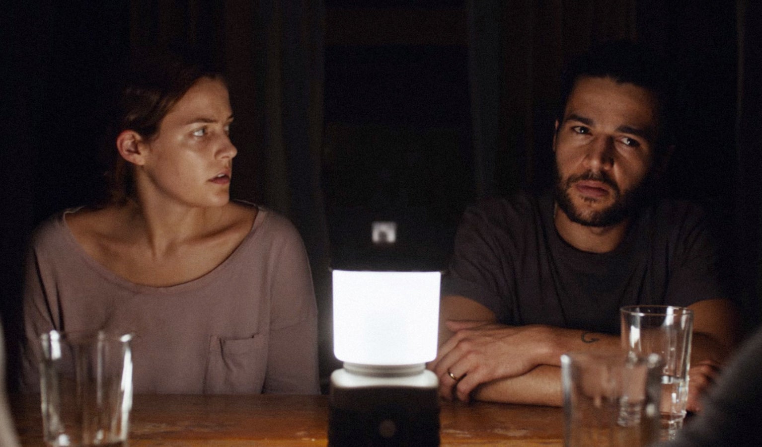 Riley Keough and Christopher Abbott sit at a table together with a camp light