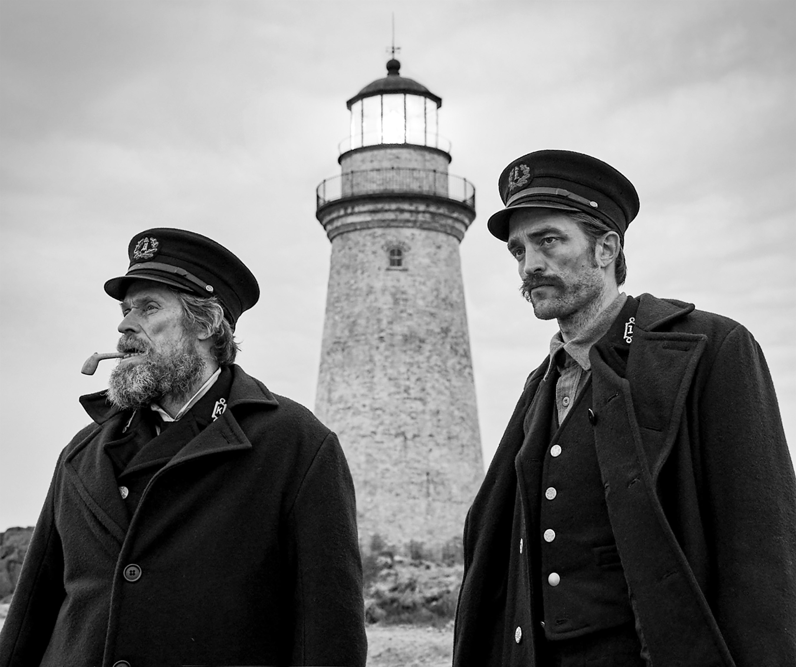 Willem Dafoe and Robert Pattionson stand in front of a lighthouse