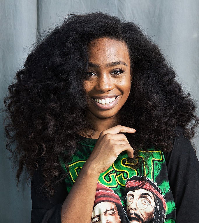 SZA smiling in a T-shirt with long curly hair parted to the side
