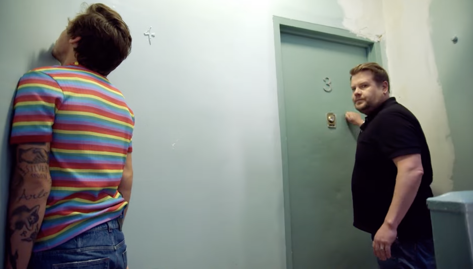 Corden knocks on an apartment door as Styles stands a few feet to the side with his face firmly planted into a wall