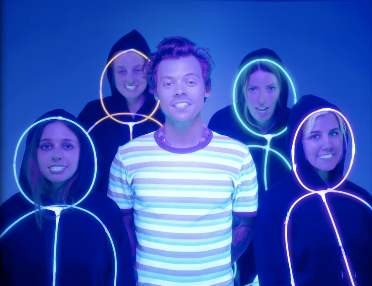 Styles stands between four women, all standing and wearing black hoodies with neon glowsticks around their faces