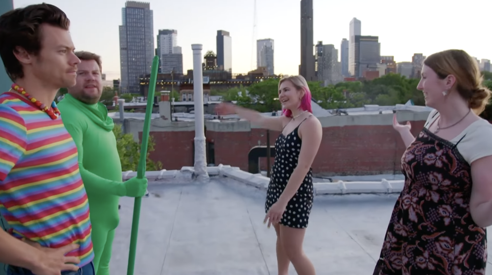 Corden wears a full-body green screen suit as Styles studies the skyline with the two women gesturing in either direction on a rooftop