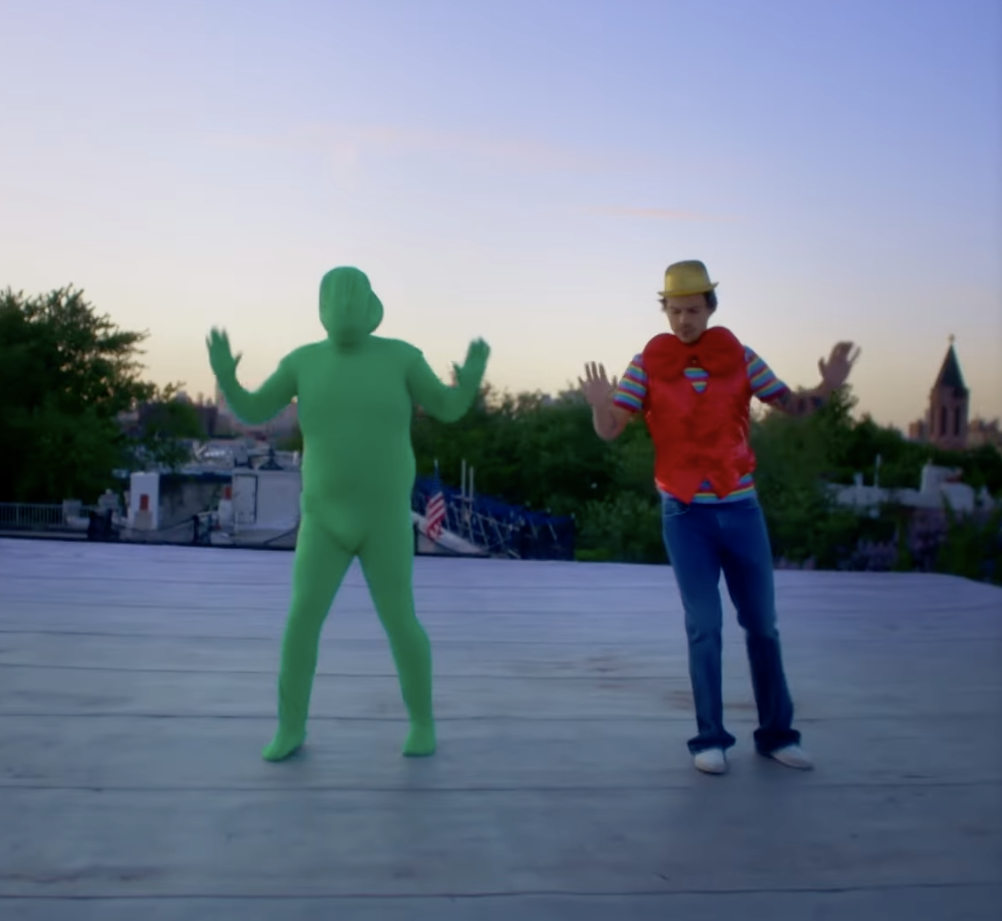 Corden in a full-body suit and Styles wearing a bowler hat and matching vest and tie stand on a rooftop with their hands up in a synchronized dance