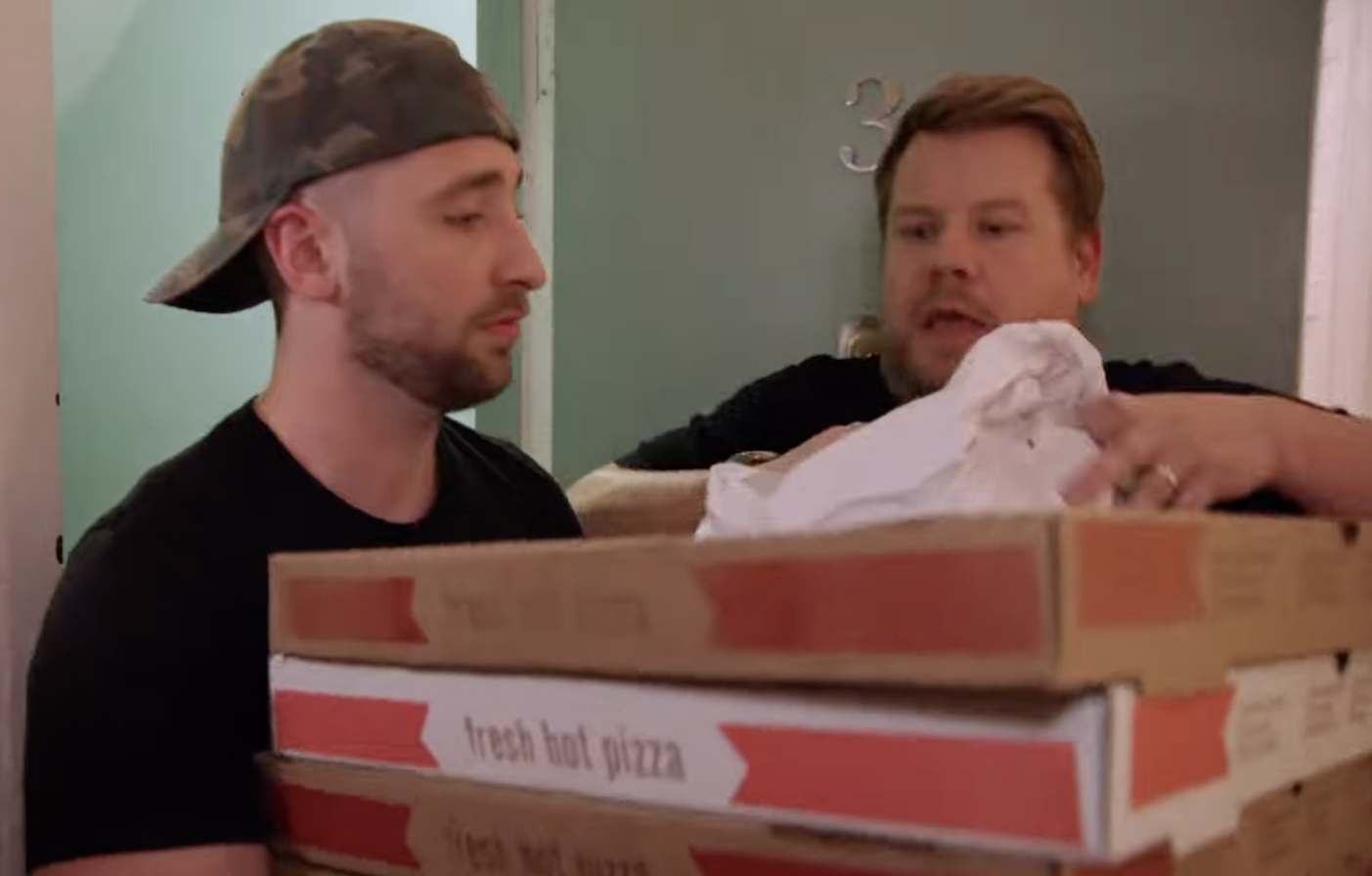 A man with a backward cap holds up multiple pizza boxes that Corden takes from him