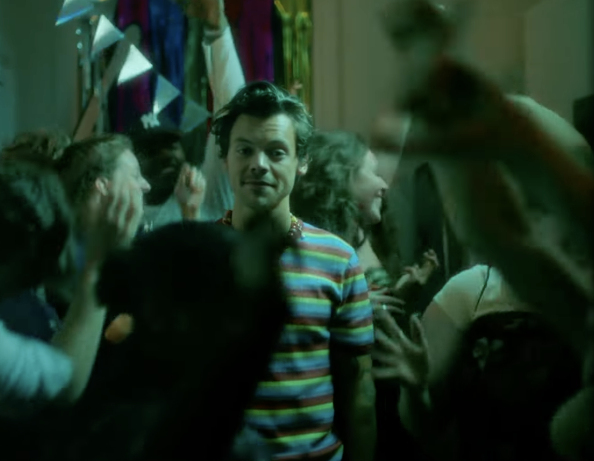 Harry stands in the middle distance, surrounded by people dancing at a party