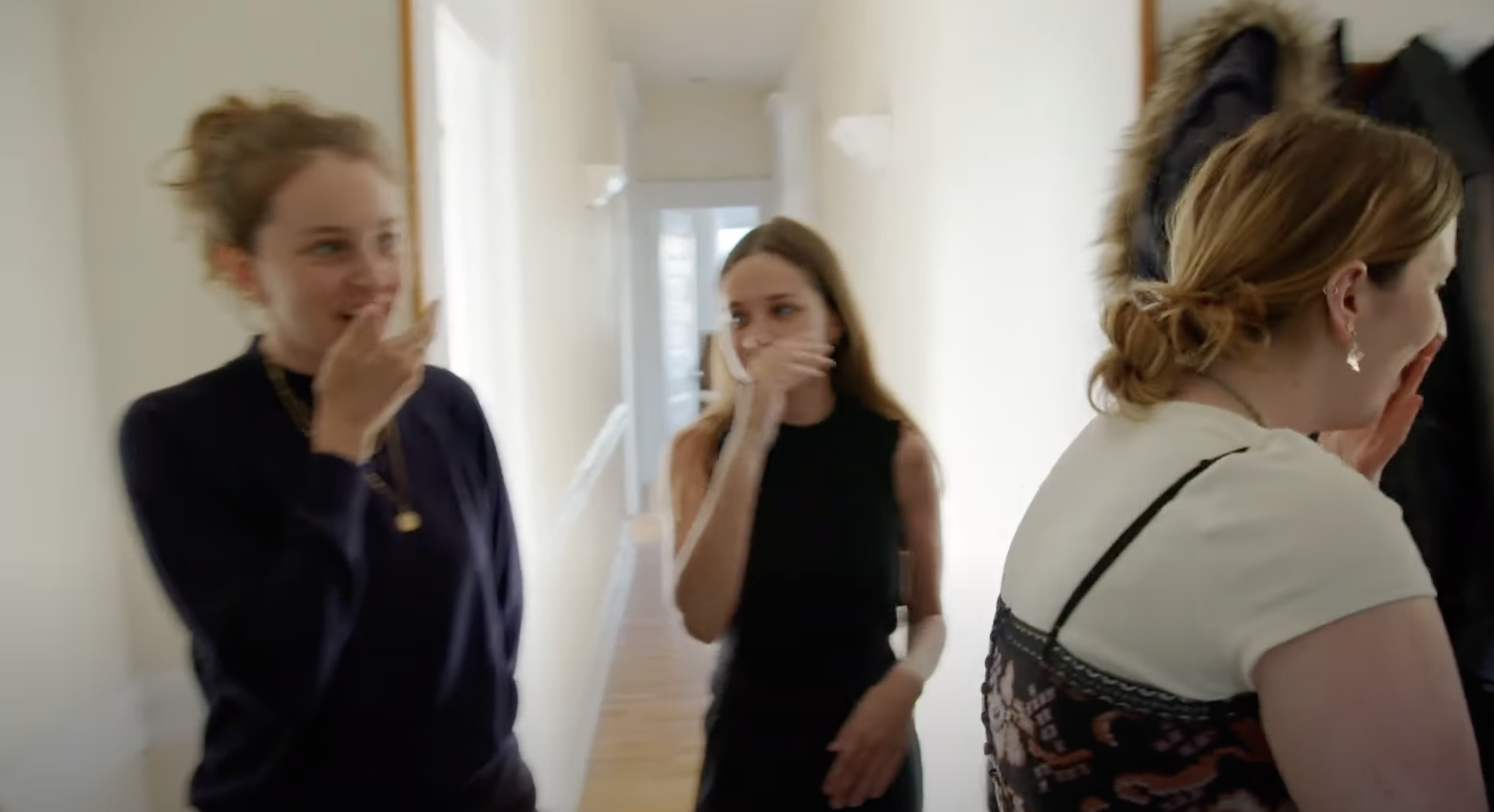 Three women stand inside their apartment, looking anxious