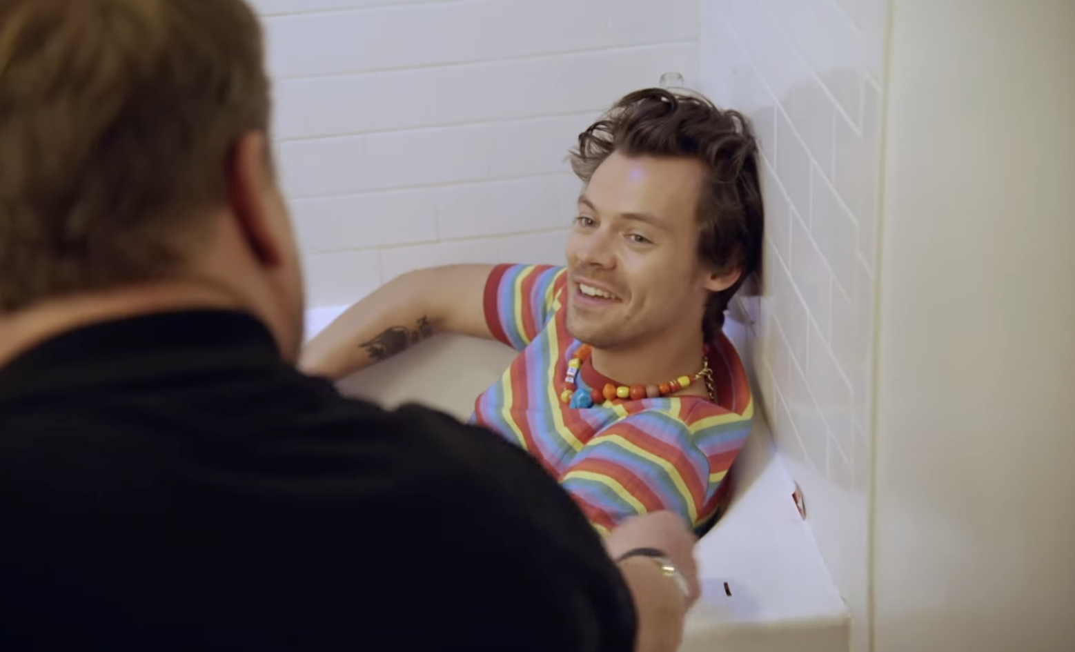 Styles sits in a bathtub, smiling at Corden