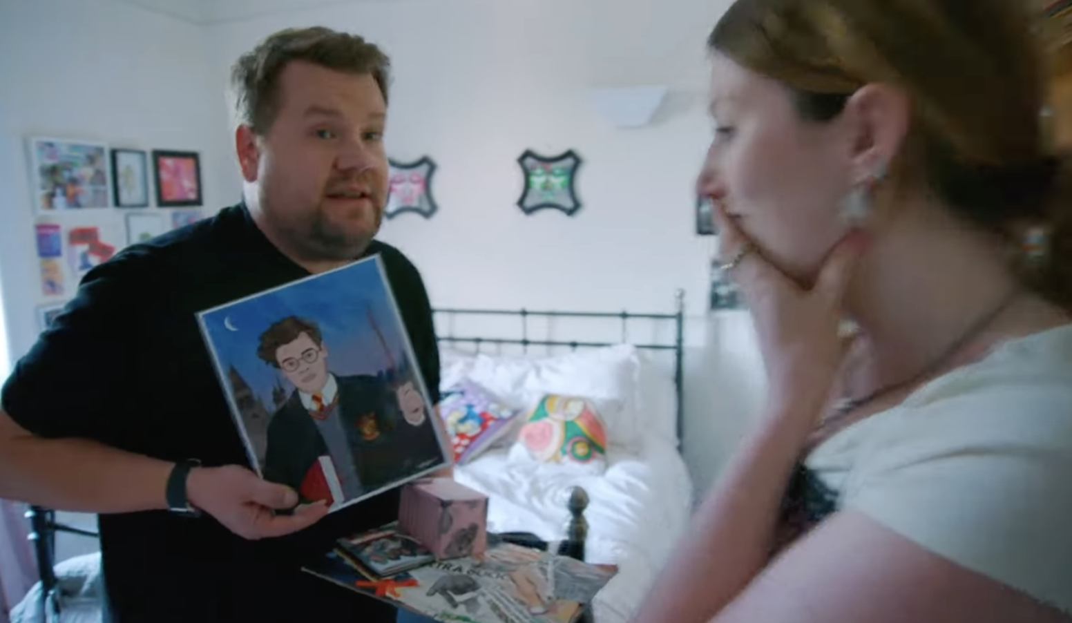 A woman holds a hand over her mouth as Corden faces her and the camera, holding up an illustration of Harry Styles as Harry Potter