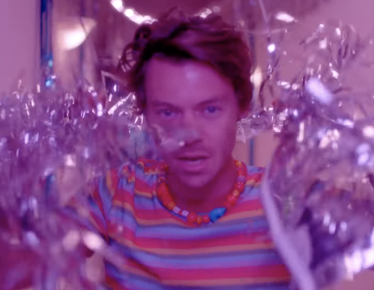 In a music video, Harry looks at the camera wearing a beaded necklace, with streamers waving on either side of his face