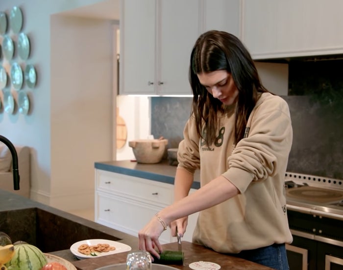 Kendall Jenner holds a cucumber on the end with her left hand, crossing over the right hand holding a knife cutting a slice