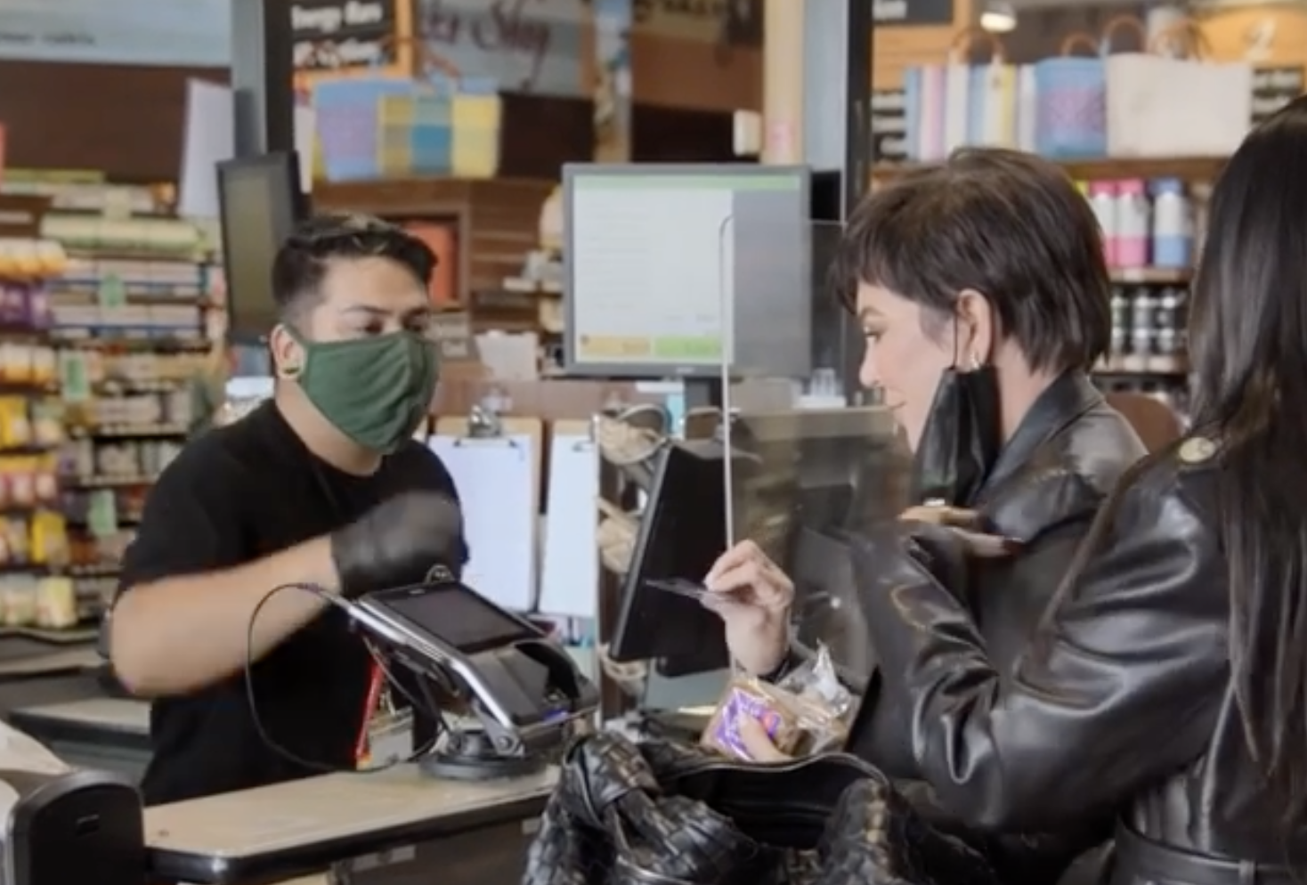 Kris Jenner, face mask off, uses a credit card at checkout by a checkout attendant wearing gloves and a face mask