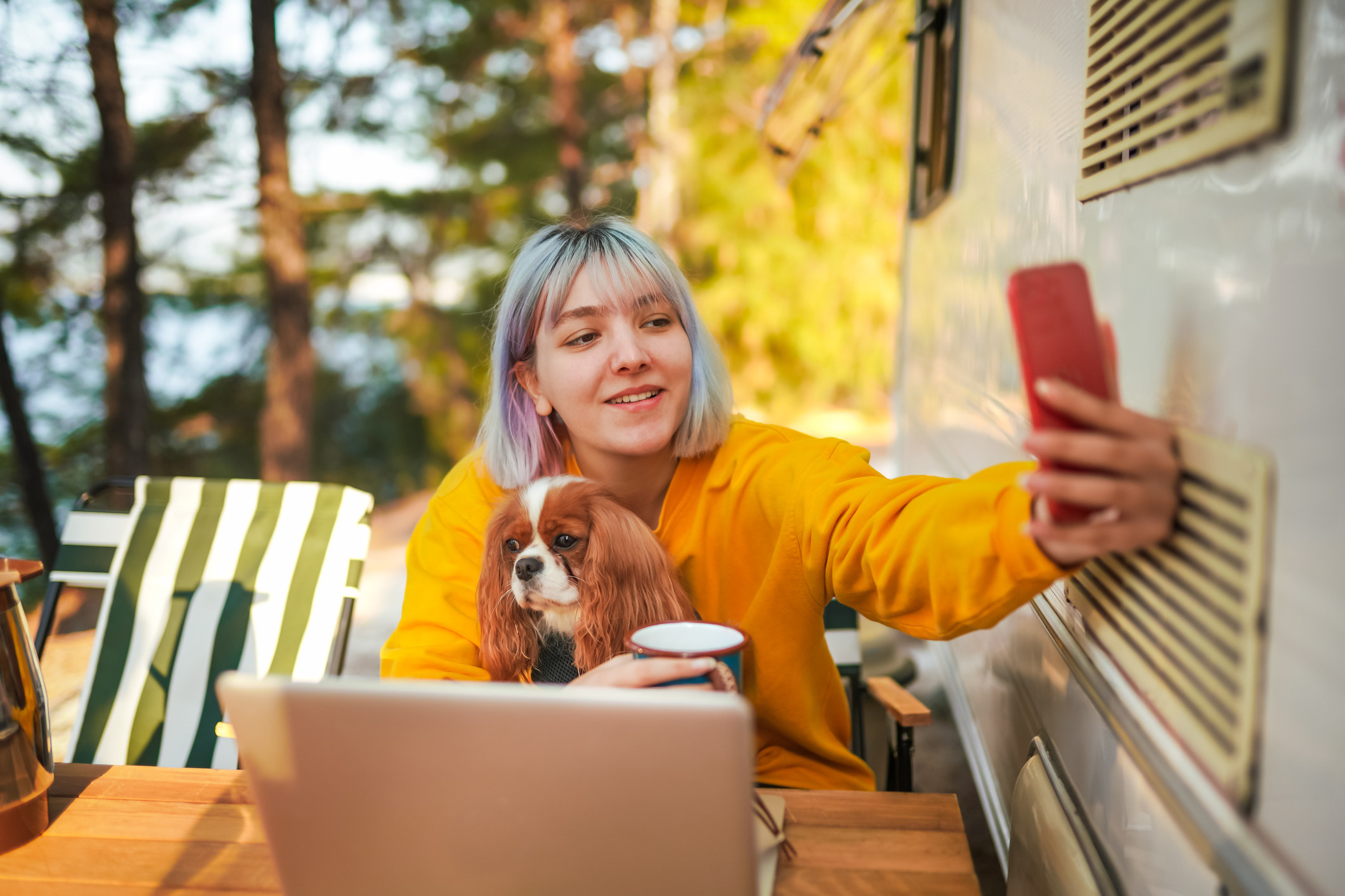 Woman sitting next to a camper with her dog. She is on her phone and computer