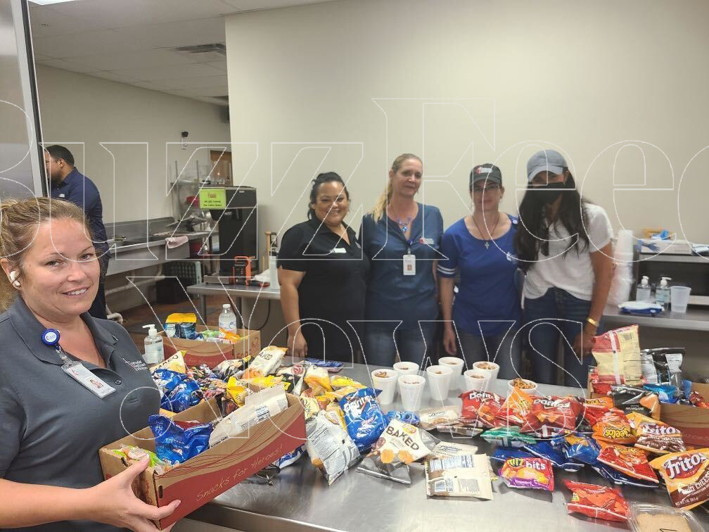 Four women stand around a snack-covered table with Meghan Markle, wearing a cap and black face mask