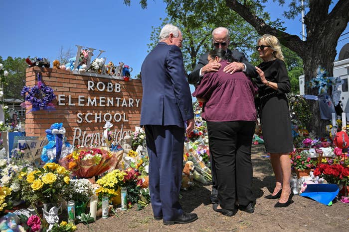 Biden and his wife stand with two other people in front of a memorial of flowers and candles in front of the "Robb Elementary School Bienvenidos" sign