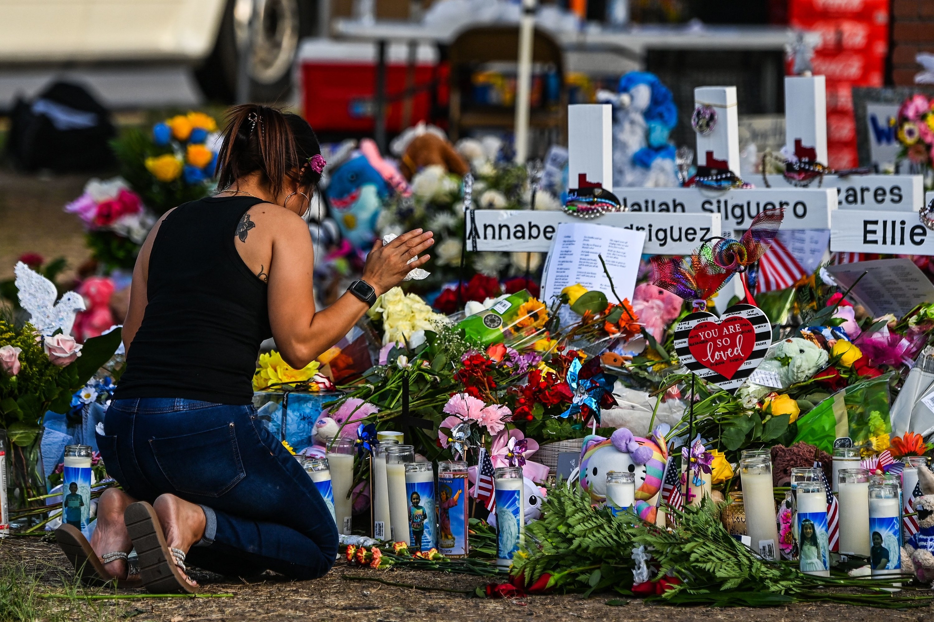 A woman kneels in front of a large arrangement of candles, flowers, and the names of victims of the shooting on crosses