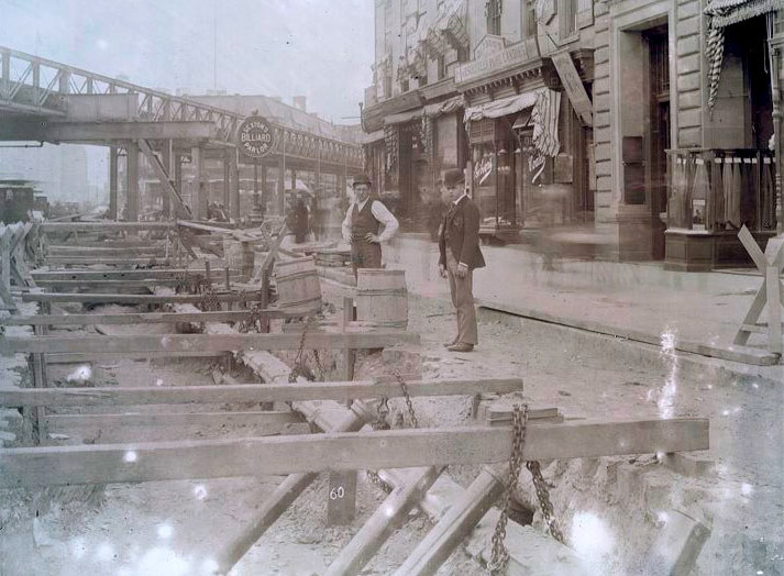 Two men stand by a street with above-ground scaffolding for subway construction