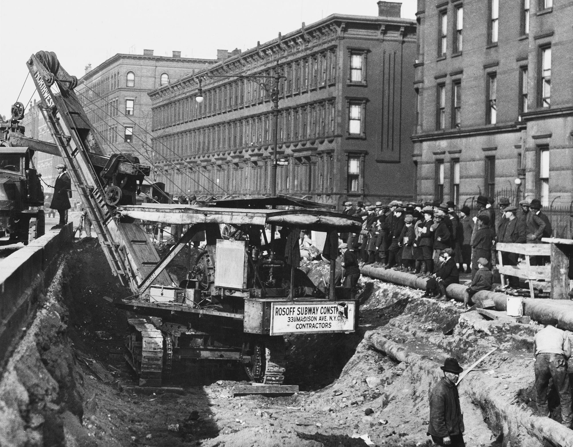 A crowd of pedestrians stand beside a large excavator machine in a hole in the street, digging a tunnel