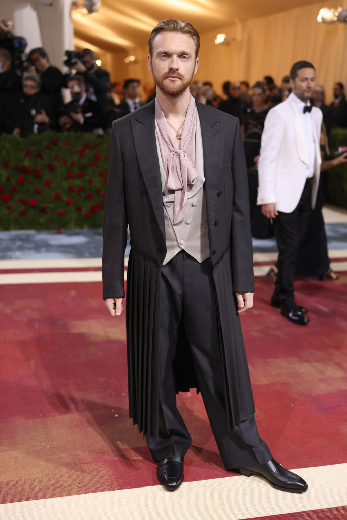 Finneas wearing a long jacket with pleats, a vest, and a scarf tie with pants