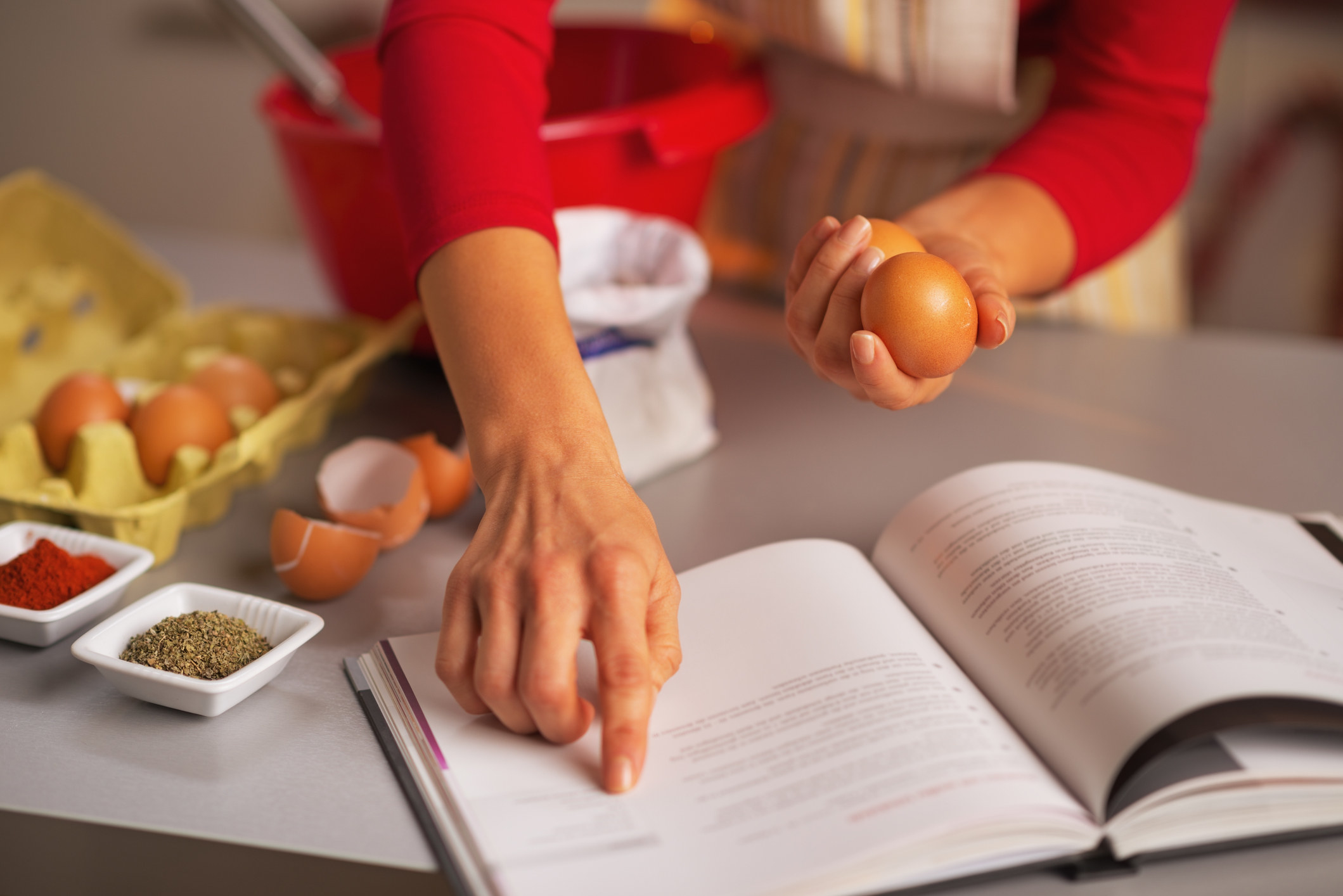A woman holding eggs and using a cookbook.