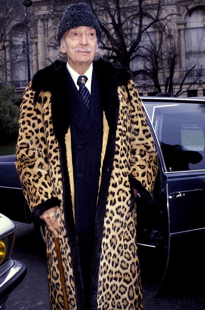a mature Dali in a long leopard print coat and cane exiting a car