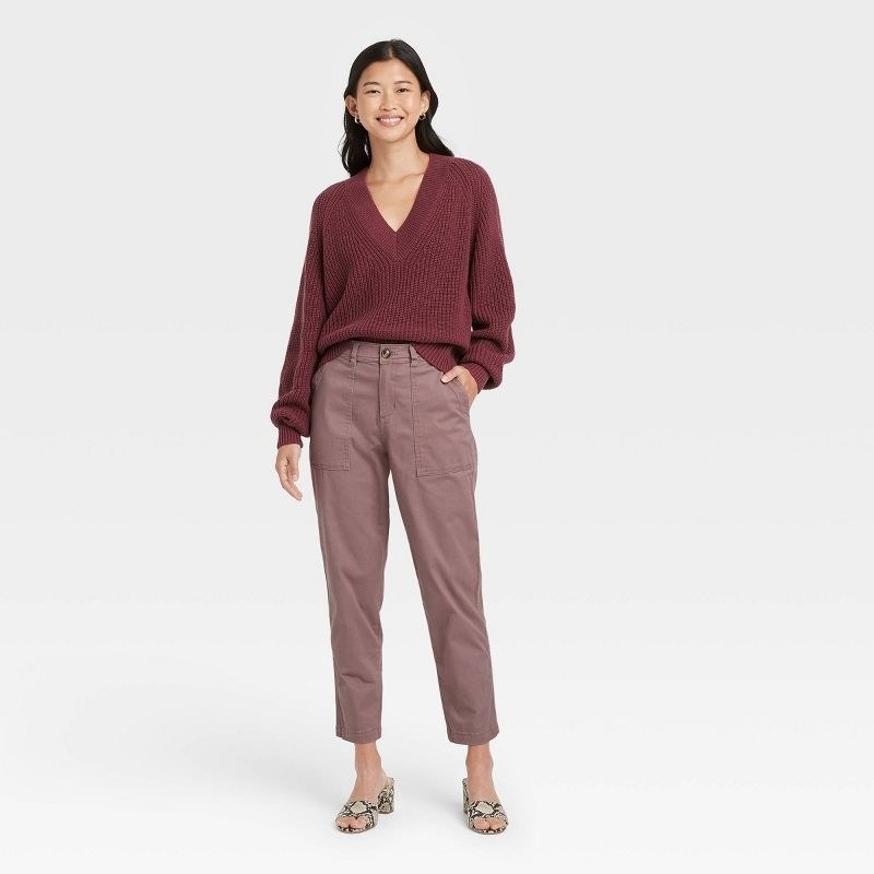 The model wears purple pants with a burgundy V-neck sweater and snake print mules