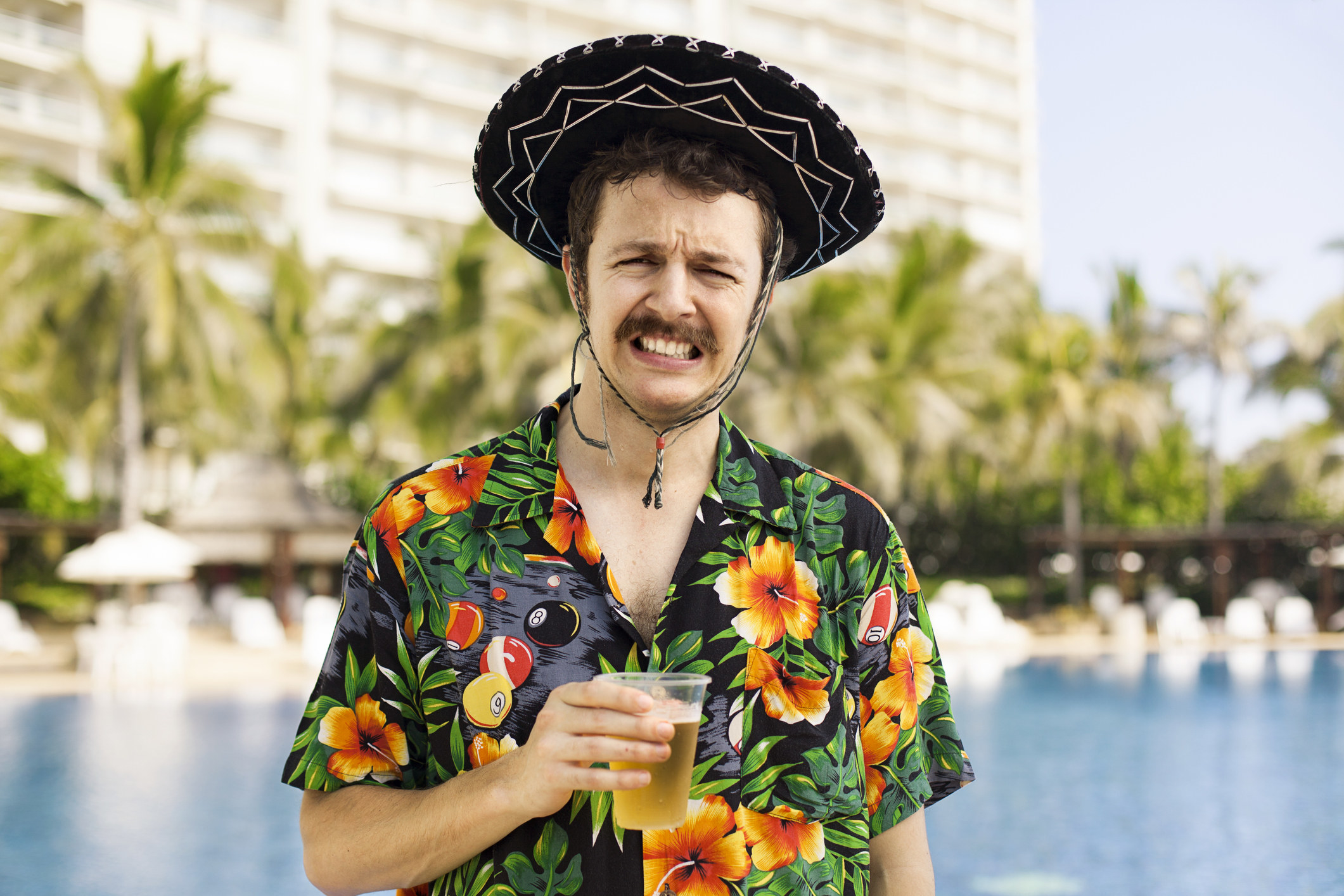 a man in a floral shirt and sombrero holding a beer and grimacing