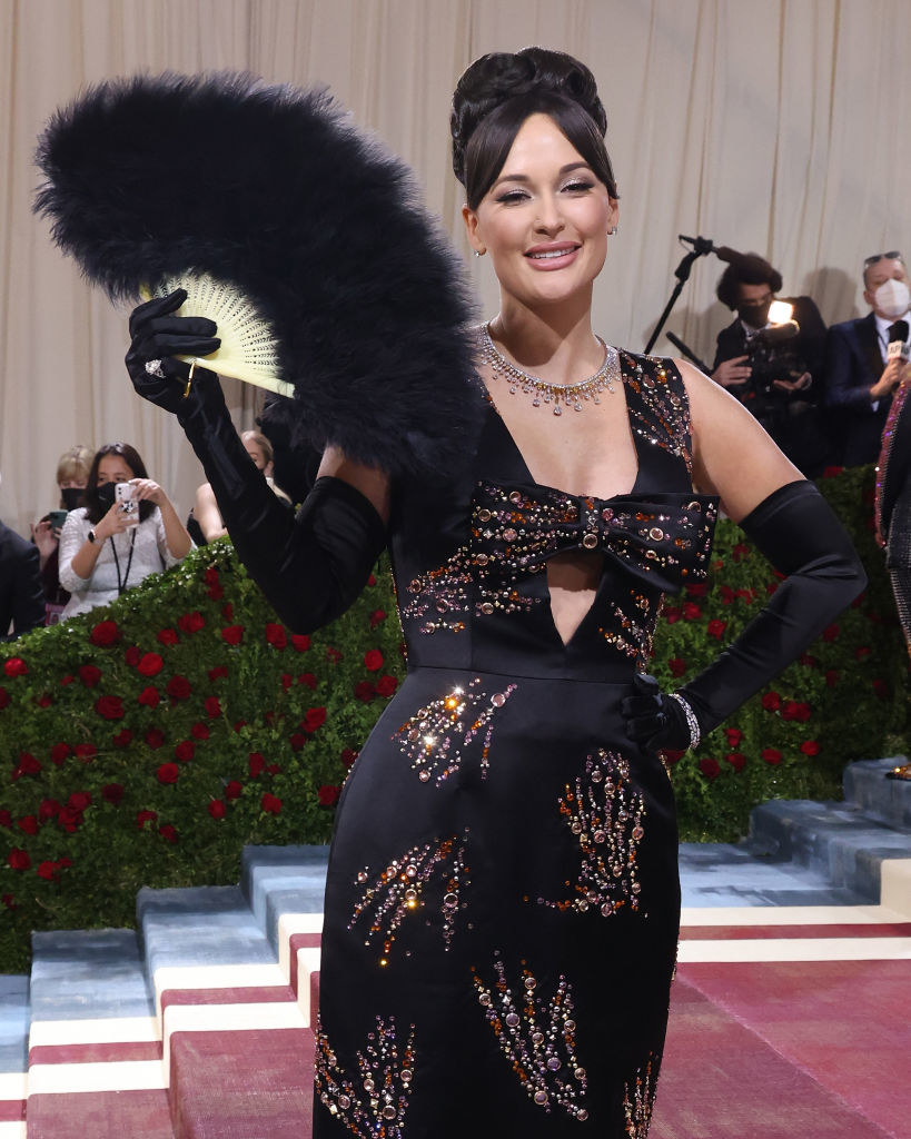 Kacey in a black dress with jewels, a bow in the middle, and holding a feathered fan