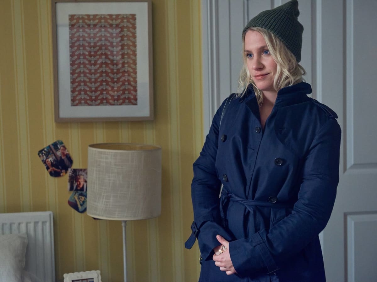 A woman, wearing a trench-coat and beanie, stands with her hands folded