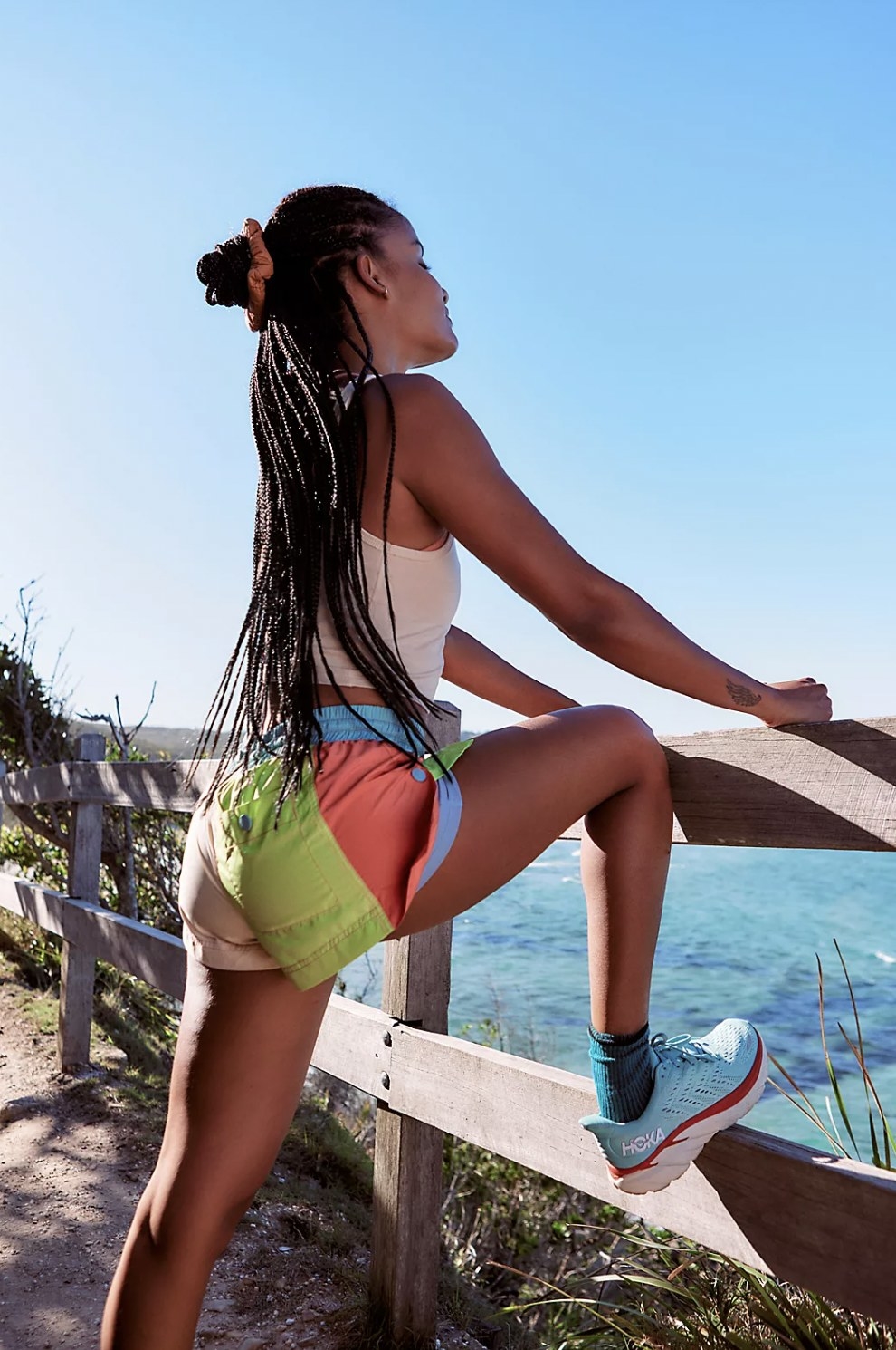 Model stretching with leg up on fence along the beach wearing green and pink colorblock shorts. Button pocket shown on backside and snap buttons on side over purple undershorts