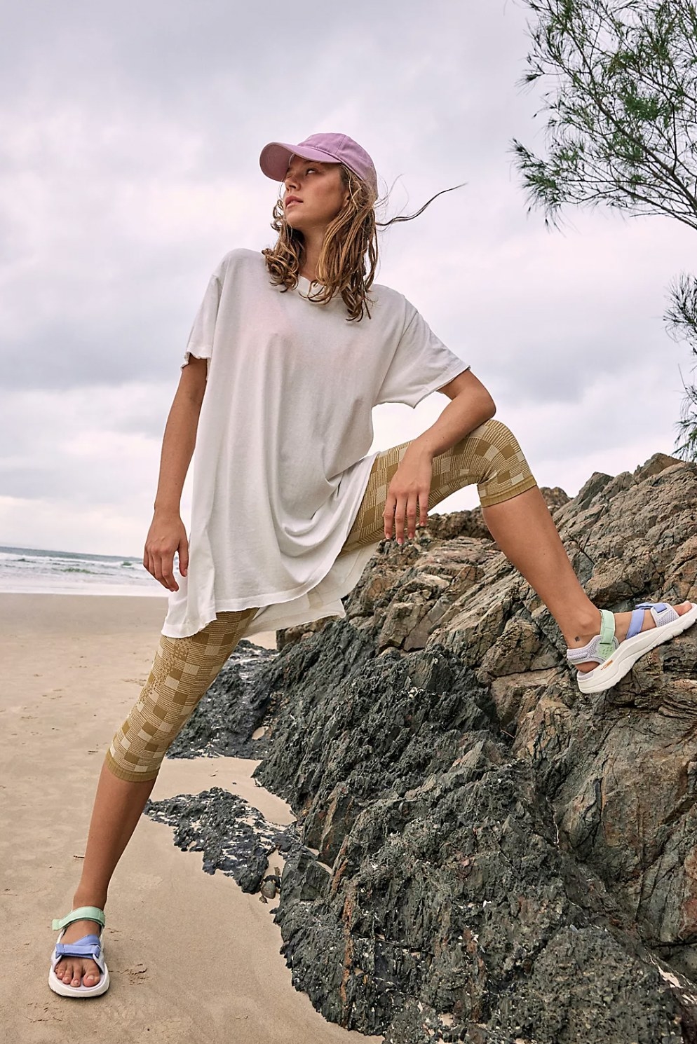 Model with leg on beach rock wearing oversized white tee with cropped leggings and sandals