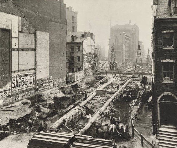 A torn-up road between rows of brick buildings show long pipes and workers for a subway construction