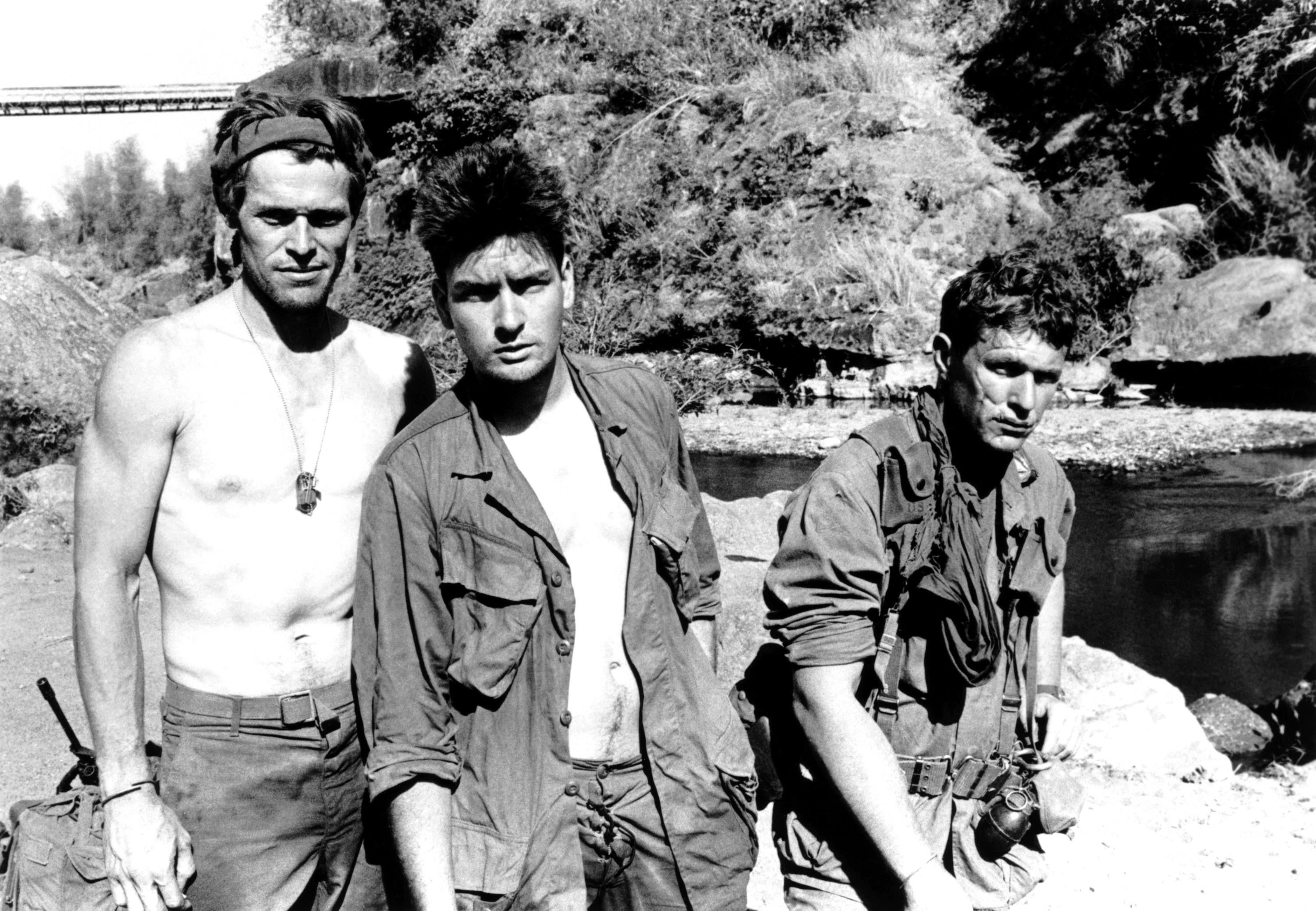 Willem Dafoe, Charlie Sheen, and Tom Berenger stand by a river