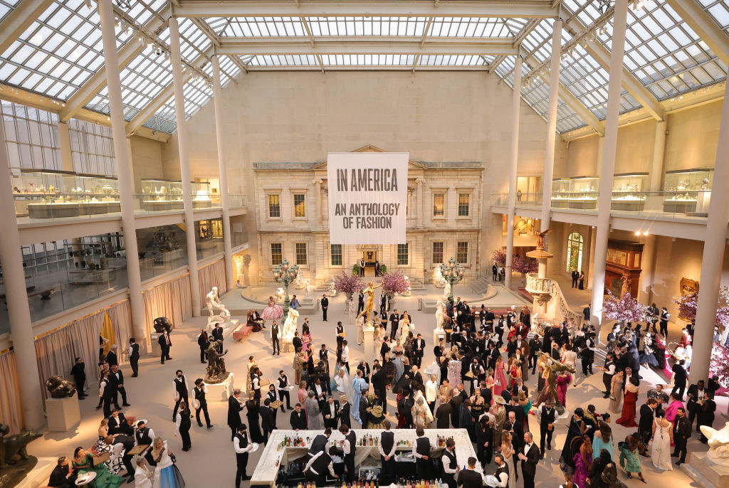 A bunch of people walking around an art exhibit with a giant title card that says &quot;In America: An Anthology of Fashion&quot;