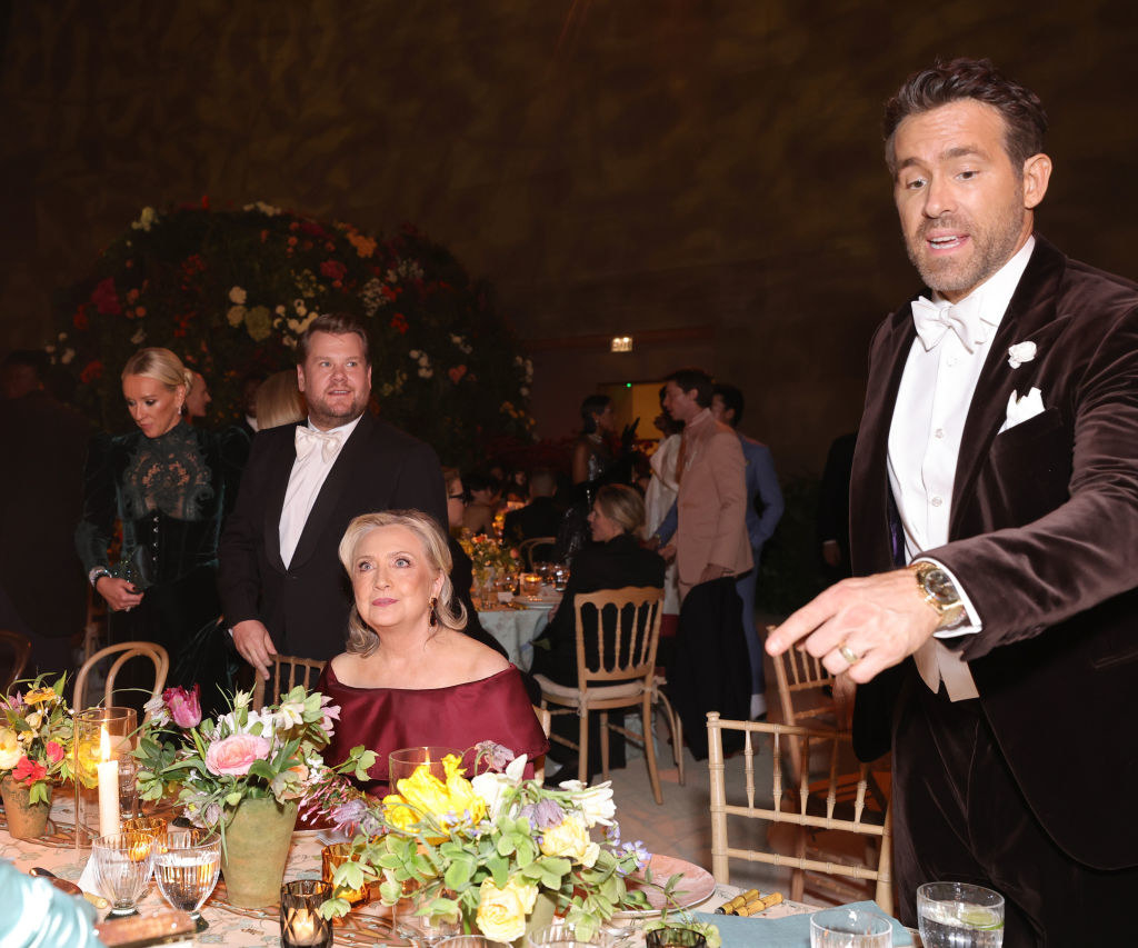 Hillary Clinton seated at a table as Ryan Reynolds talks to someone nearby