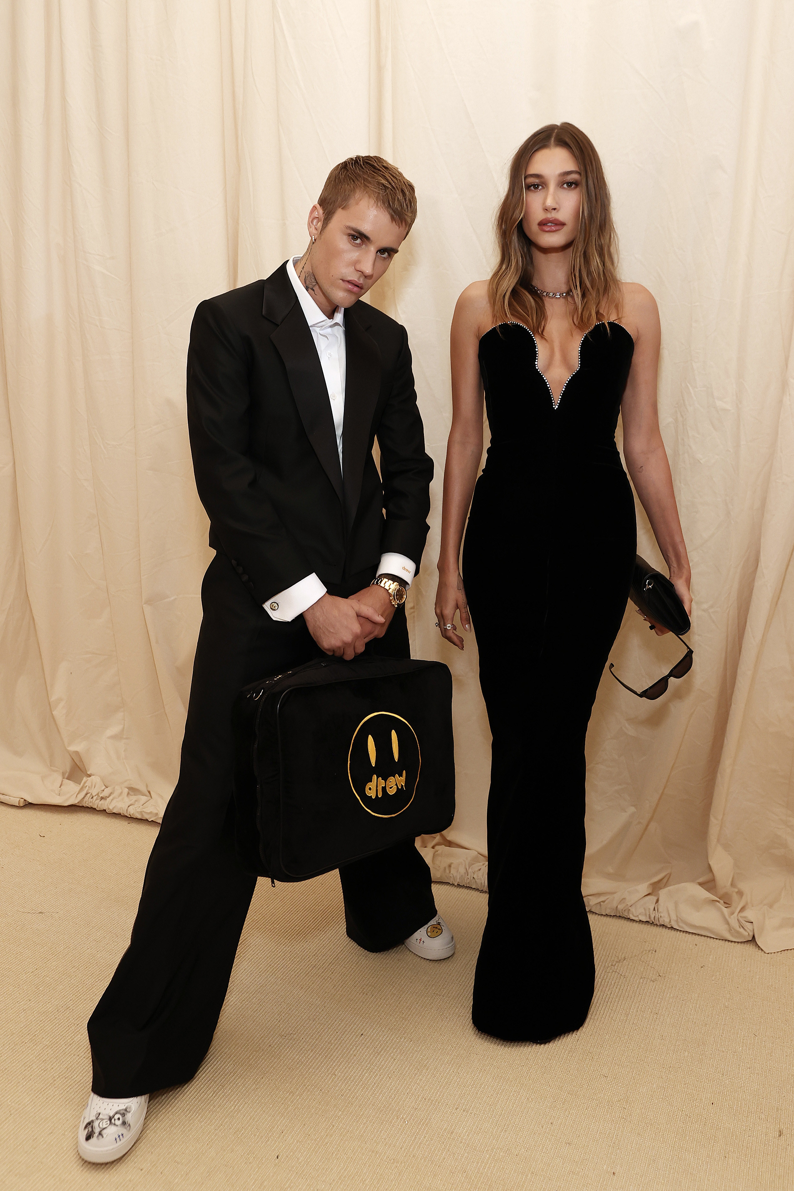 Justin, in a suit. posing as he holds onto a bag and stands by Hailey who is wearing a strapless dress