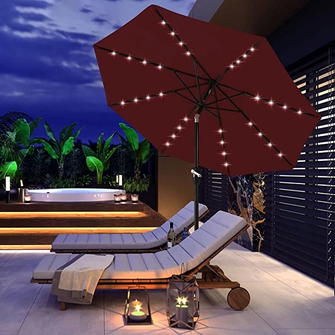 An umbrella tilted up beside lounge chairs with the lights on it at night