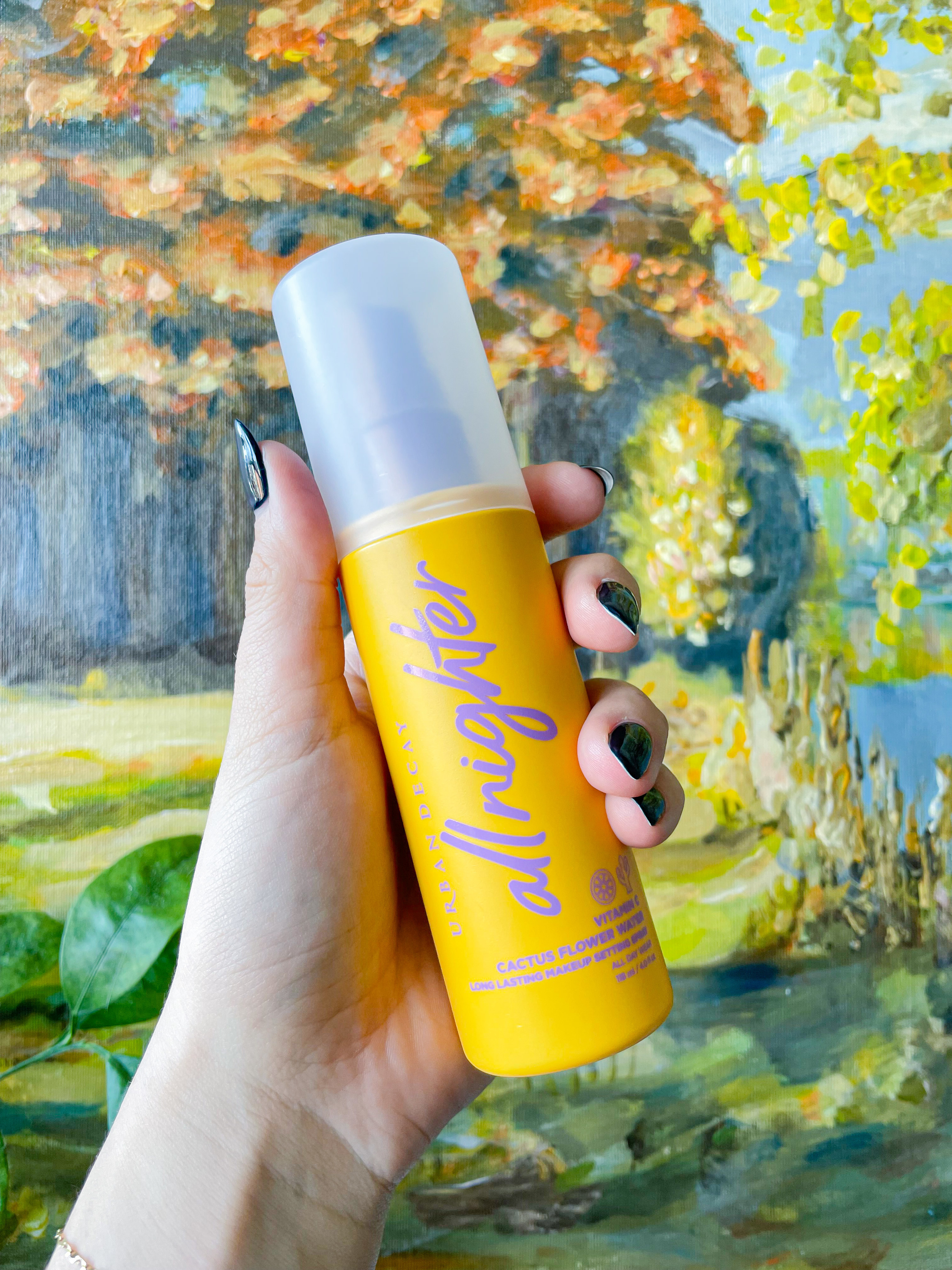Victoria holding up a bottle of the vitamin c-infused setting spray against a painting