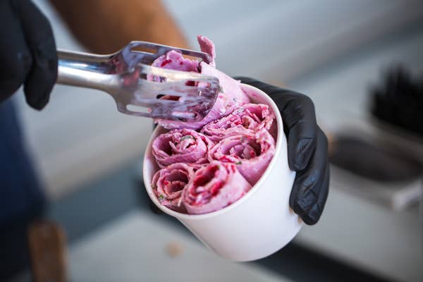 Pink ice cream has been flattened and rolled into spirals, then is put into a paper cup with tongs