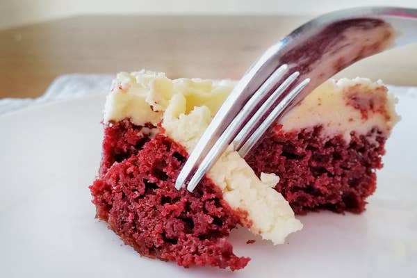 A close up shot of a piece of bright red cake with white icing being picked up with a fork