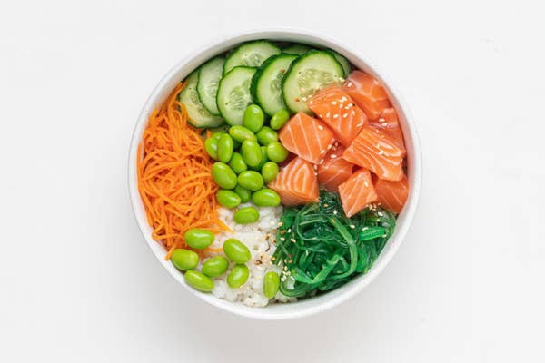 A white bowl containing raw salmon, cucumber, edamame, carrot, rice, and seaweed